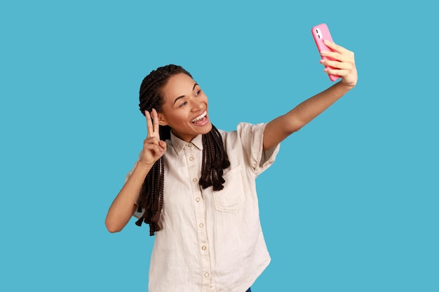 La donna felice con il sorriso a trentadue denti fa il segno di pace o il gesto V alla fotocamera del telefono cellulare pone per fare selfie o videochiamate indossando una camicia bianca Scatto in studio al coperto isolato su sfondo blu