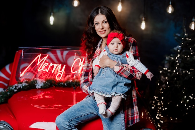 La donna felice con una bambina con un fiocco rosso sulla testa è seduta e si diverte su un'auto retrò in studio di Natale.