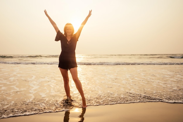 La donna felice del viaggiatore in vestito corto nero si gode la sua vacanza sulla spiaggia tropicale al tramonto romantico silhouette estate tempo primaverile mani in alto piacere spf e crema solare