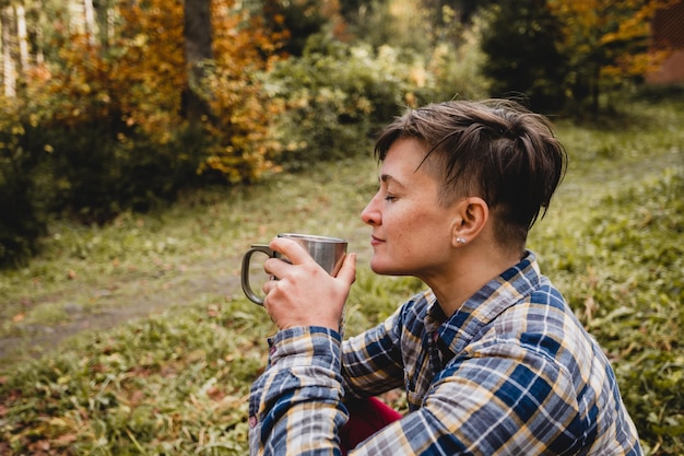 La donna felice è seduta nella foresta autunnale con gli occhi chiusi Sta tenendo una tazza di bevanda calda