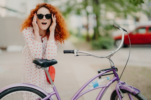 La donna felice ha i capelli ricci e ricci, si appoggia alla sella della sua bicicletta,