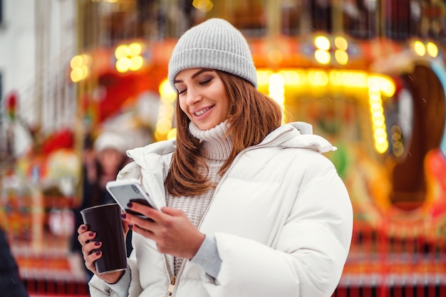 La donna felice indossa in piumini bianchi legge lo smartphone nella città di natale mentre tiene il caffè