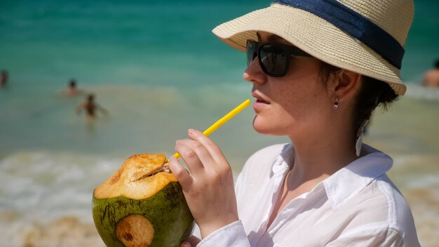La donna felice si siede sulla spiaggia e beve un cocktail di cocco