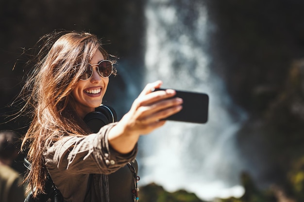 La donna felice si sta godendo il paesaggio in montagna facendo selfie accanto alla bellissima cascata all'aperto.