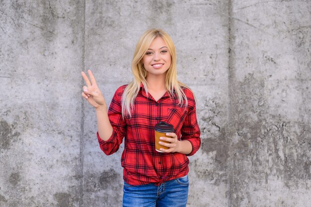 La donna felice sorridente con capelli biondi vestita in abiti casual sta mostrando il segno di vittoria e sta tenendo il caffè del mattino