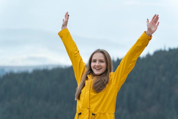 La donna felice sta con le mani sollevate sullo sfondo della foresta Ragazza turistica in impermeabile giallo in montagna