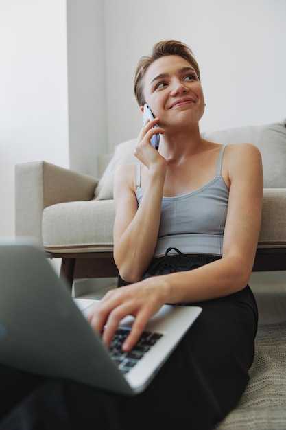 La donna freelance con laptop e telefono lavora da casa seduta sul pavimento nei suoi vestiti di casa con uno spazio di copia gratuito per il taglio di capelli corto