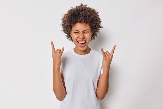 La donna gioiosa spensierata con i capelli ricci fa il gesto del rock'n'roll heavy metal segno gode della musica preferita vestita casualmente isolata su sfondo bianco trascorre il tempo libero alla festa. Linguaggio del corpo