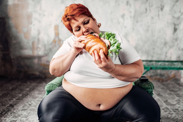 La donna grassa si siede sulla sedia e mangia il panino, bulimico