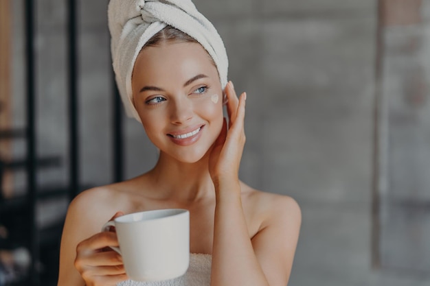 La donna graziosa applica il trucco minimo della crema per il viso avvolto in un asciugamano da bagno gode del caffè a casa Concetto di rilassamento di bellezza