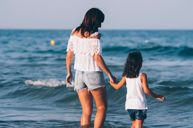 La donna graziosa con i jeans corti e sua figlia che posano sull'acqua della spiaggia