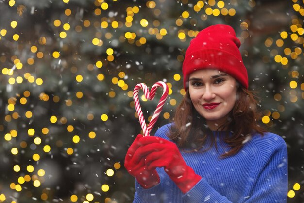 La donna graziosa indossa la caramella della tenuta del berretto rosso all'albero di Natale. Spazio vuoto