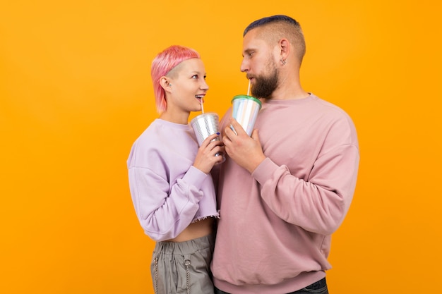 La donna graziosa insolita con i capelli e il tatuaggio rosa corti beve il caffè e si diverte con il suo ragazzo isolato su fondo arancio