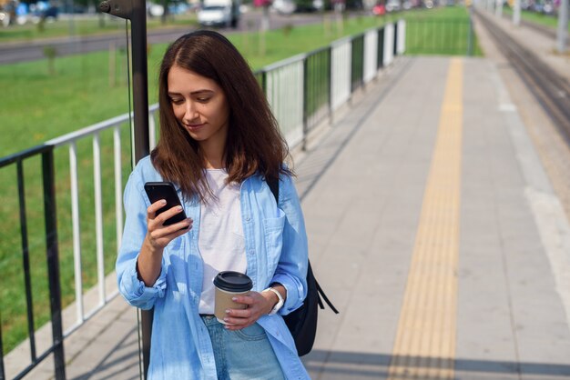 La donna graziosa usa lo Smart Phone e beve il caffè di mattina per andare mentre aspetta il tram o l'autobus sulla stazione pubblica.