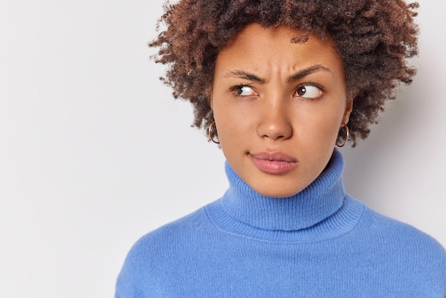 la donna guarda attentamente da parte ha un'espressione sospettosa vestita con un dolcevita blu casual isolato su bianco con uno spazio vuoto per la pubblicità