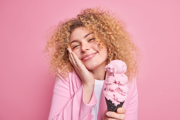 la donna guarda con gioia la telecamera tiene la mano sulla guancia sorride delicatamente tiene un gustoso gelato grande in cialda vestita con una giacca elegante