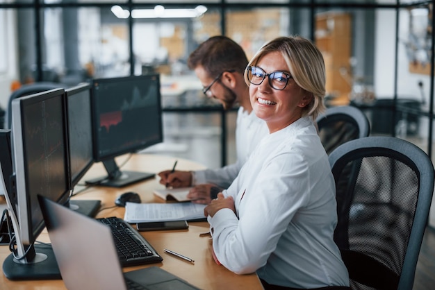 La donna guarda nella telecamera. Due agenti di cambio in abiti formali lavorano in ufficio con il mercato finanziario.