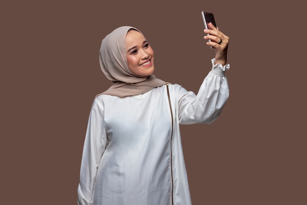 La donna HIjab si fa un selfie usando uno smartphone isolato su uno sfondo chiaro