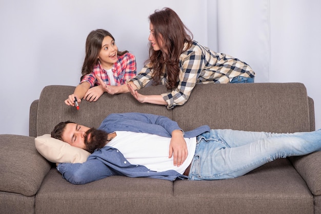 La donna impedisce al bambino cattivo di applicare il rossetto all'uomo che dorme sul divano a casa, in famiglia.