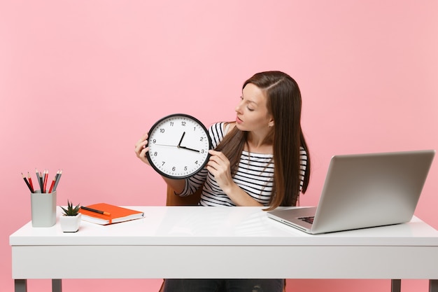 La donna in abiti casual tiene a guardare la sveglia rotonda mentre è seduta a lavorare alla scrivania bianca con un laptop pc contemporaneo isolato su sfondo rosa pastello. Concetto di carriera aziendale di successo. Copia spazio.