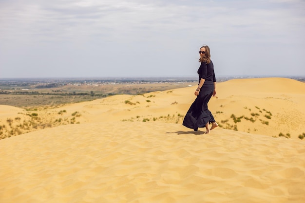 La donna in abito lungo nero cammina attraverso le dune del deserto in estate con la schiena