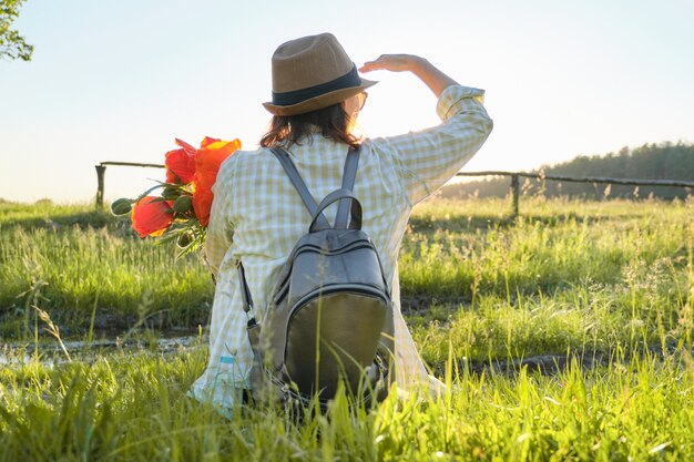 La donna in cappello con i fiori dello zaino che si siedono indietro sull'erba che gode della natura di estate