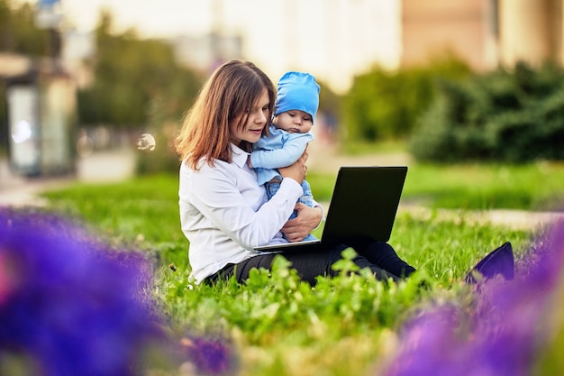 La donna in congedo di maternità lavora da remoto online fuori si siede davanti al laptop con il bambino in braccio