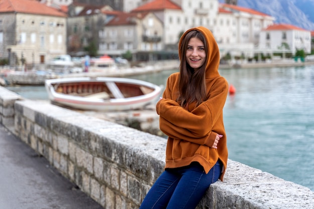 La donna in felpa con cappuccio arancione fa una pausa il mare in città Perast sui precedenti nel Montenegro