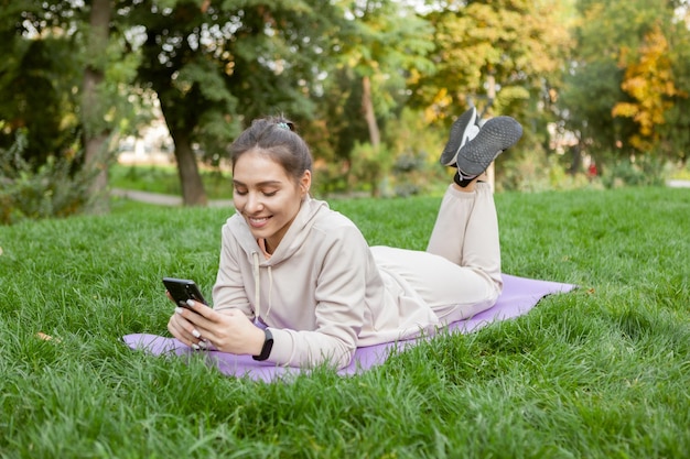 La donna in forma ha tempo di riposo sdraiata sul tappetino e utilizza lo smartphone nel parco Fitness o yoga all'aperto