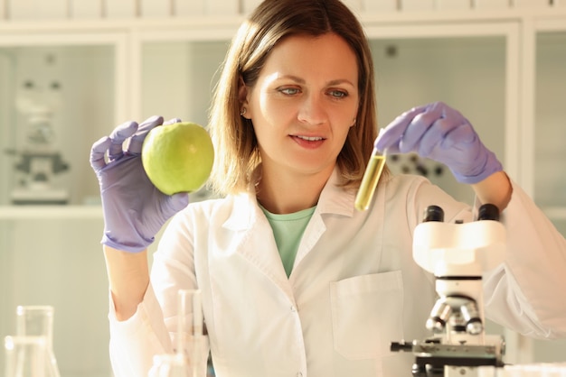 La donna in guanti di gomma tiene una mela fresca e un tubo con un campione di liquido giallo nello specialista delle mani