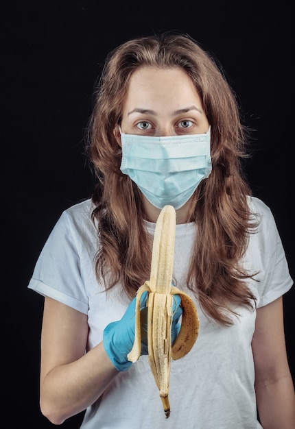 La donna in guanti e maschera medica protettiva cerca di mangiare una banana sul muro nero