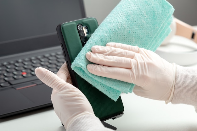 La donna in guanti pulisce il phomelaptop con tessuto umido e disinfettante durante il covid 19. Disinfezione phome e tastiera del laptop con disinfettante alcolico da parte di una donna in maschera che si illumina sul posto di lavoro, scrivania.