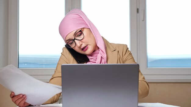 La donna in hijab esamina i documenti e lavora al laptop mentre parla al telefono