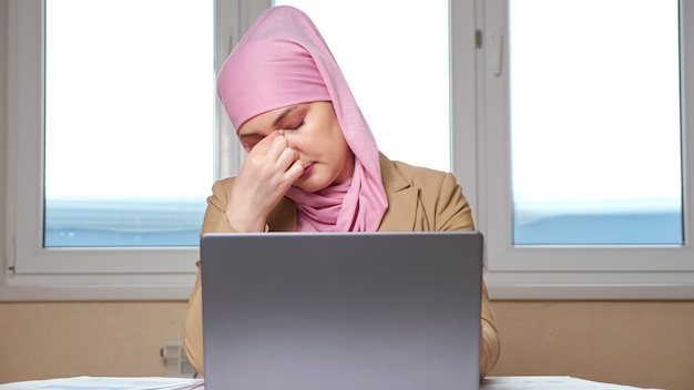 La donna in hijab si tiene la testa e massaggia le tempie mentre lavora su un laptop.