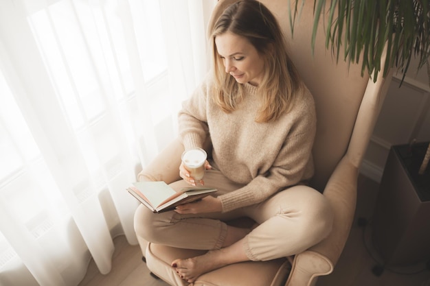 La donna in maglione lavorato a maglia caldo beige legge il libro a casa vicino alla finestra