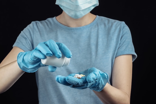 La donna in maschera facciale e guanti medicali tiene una bottiglia di pillole sul muro nero.