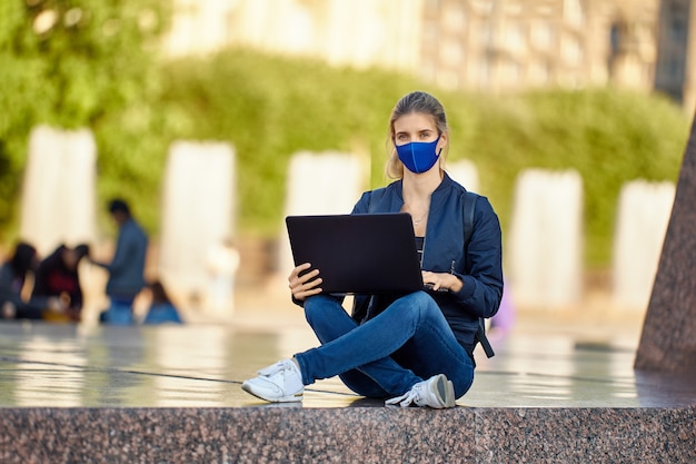 La donna in maschera protettiva facciale con il computer portatile si siede all'aperto