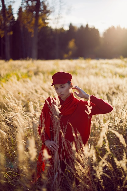 La donna in un berretto e sciarpa rossi del maglione sta in un campo in autunno nell'erba secca alta