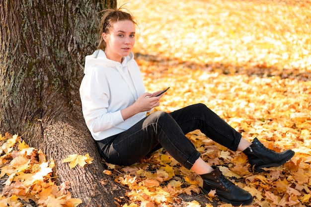 La donna in un maglione con cappuccio bianco con cappuccio si siede a terra nel parco e tiene in mano un cellulare. ragazza carina usa lo smartphone per la comunicazione Femmina che invia un messaggio a un messaggio Concetto di comunicazione