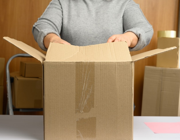 La donna in un maglione grigio sta imballando scatole di cartone marroni su un tavolo bianco, dietro una pila di scatole. Concetto in movimento