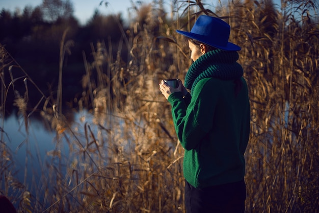 La donna in un maglione lavorato a maglia verde e un cappello blu sta vicino ai cespugli al tramonto e beve il tè da una tazza di termos di ferro