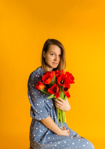 La donna in un vestito blu si siede con un mazzo di tulipani rossi su una parete gialla