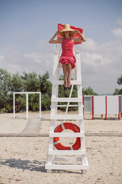 La donna in un vestito rosso si siede sulla stazione di salvataggio con una bandiera e un salvagente sulla spiaggia sabbiosa del mare