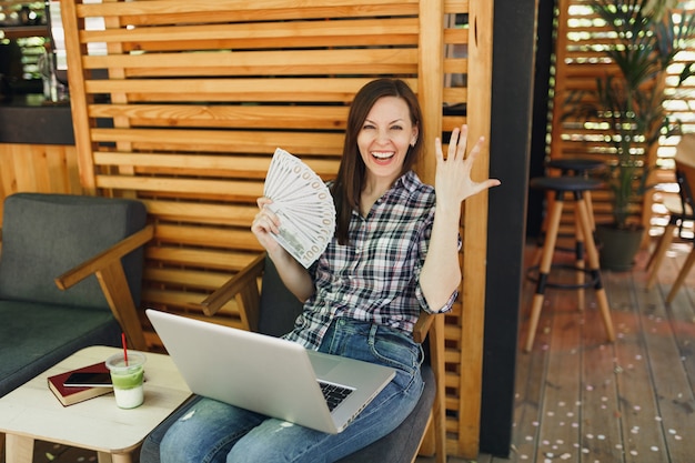 La donna in una caffetteria all'aperto di strada seduta con un moderno computer portatile, tiene in mano un mucchio di banconote in dollari, denaro contante
