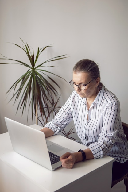 La donna in una camicia a righe bianca invecchiata siede al computer a casa