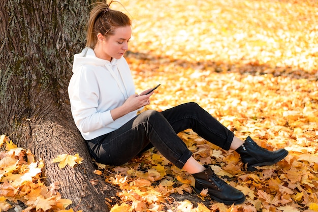 La donna in una felpa con cappuccio bianca con cappuccio si siede a terra nel parco e tiene un cellulare tra le mani.