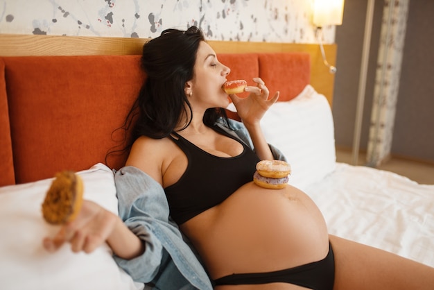 La donna incinta con la pancia mangia le torte dolci a letto a casa. Gravidanza, gola nel periodo prenatale. Mamma in attesa che riposa in camera da letto