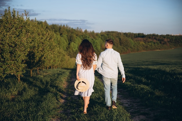 La donna incinta felice alla moda ed alla moda e suo marito hanno vestito un tono bianco e blu pastello nel campo sul tramonto