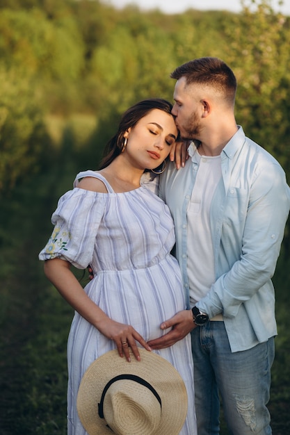 La donna incinta felice alla moda ed alla moda e suo marito hanno vestito un tono bianco e blu pastello nel giardino sul tramonto