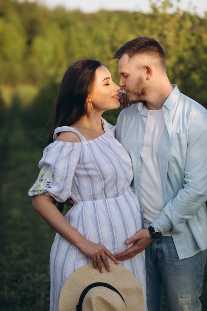 La donna incinta felice alla moda ed alla moda e suo marito hanno vestito un tono bianco e blu pastello nel giardino sul tramonto